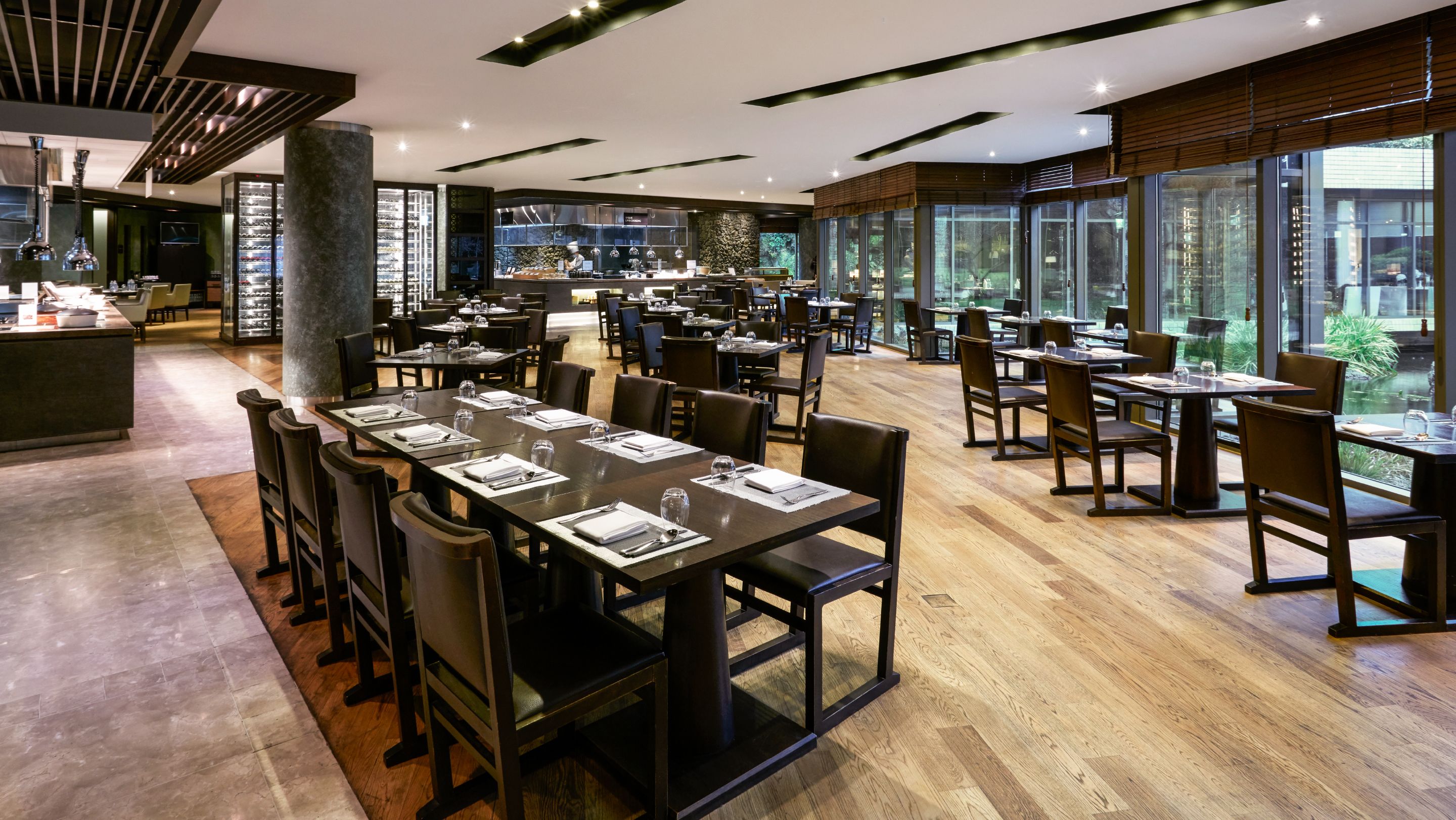 A modern restaurant with long tables and chairs, a bar area, and large windows offering a view of the outdoors.