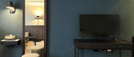 A hotel room with a large mirror, a flat-screen TV on a desk, a lamp, a small table with a tissue box, and a round ottoman.