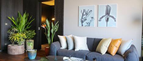 A modern living room with a grey sofa, multiple cushions, a coffee table, and several potted plants.