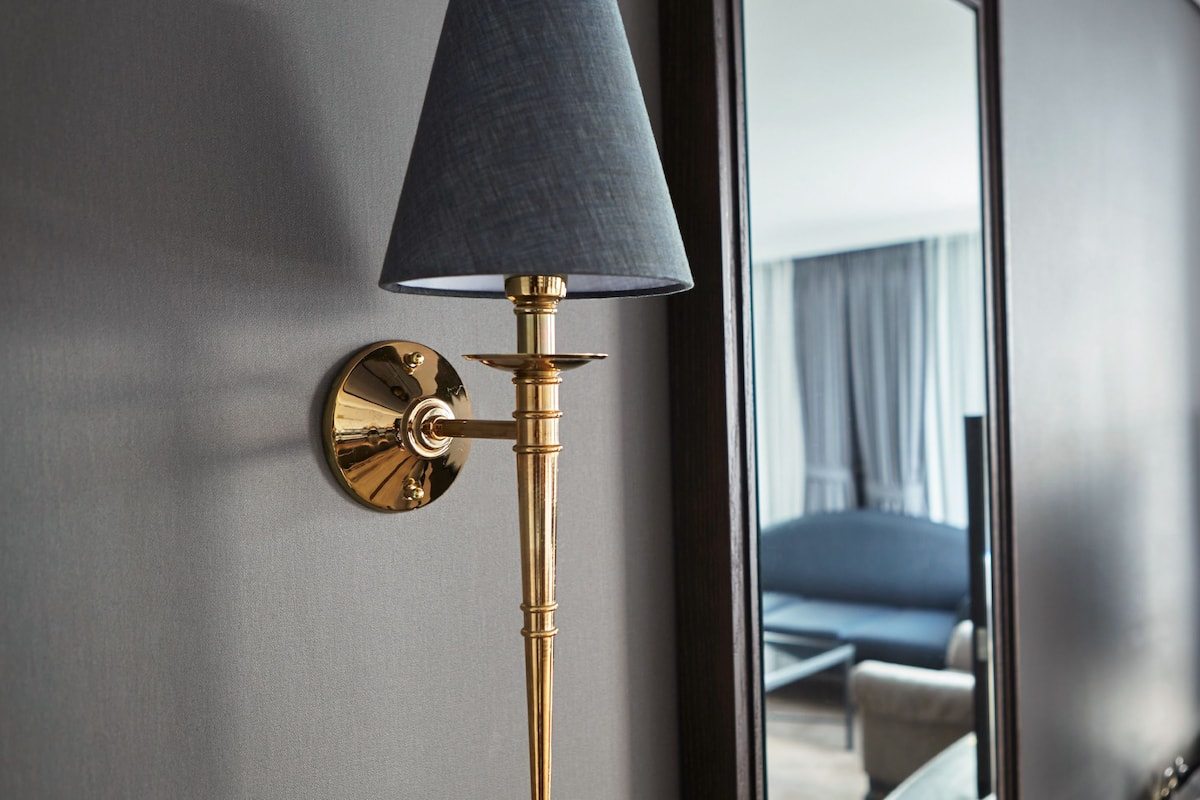 Een moderne kamer met een wandlamp, een grote spiegel die een bed en een televisie reflecteert, en een raam met gordijnen.