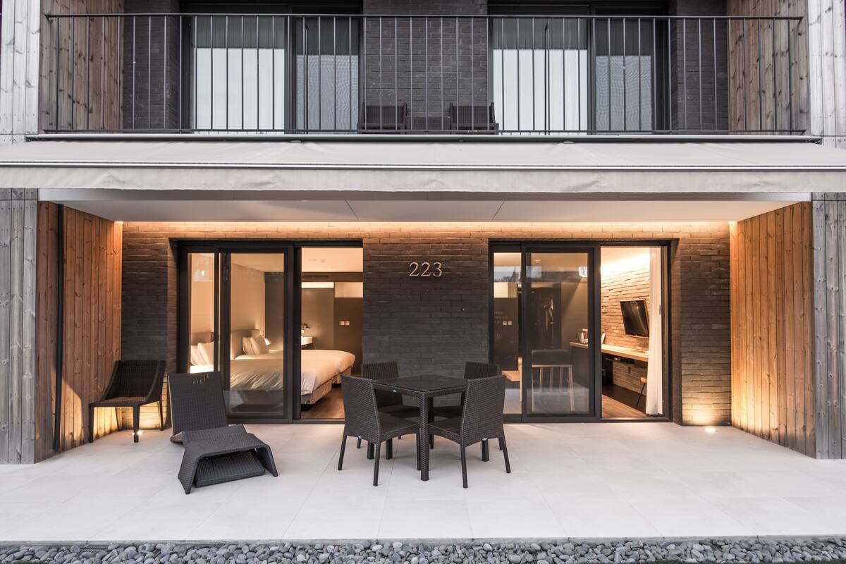 Moderne hotelentree met het cijfer '223' boven de deur, een overdekt terras met stoelen, en een zichtbare slaapkamer met bed en televisie.