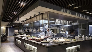 Una cocina moderna con chefs preparando comida, un bufé con diversos platos y un diseño limpio y contemporáneo.