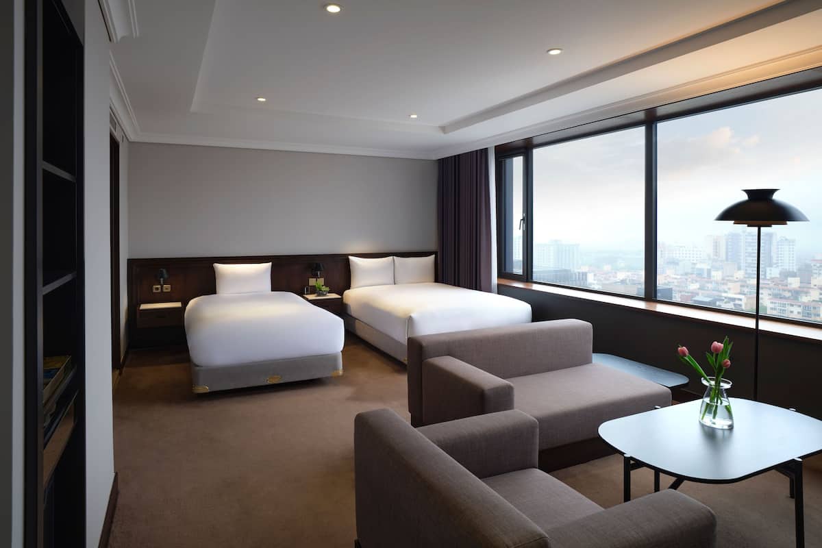 Een hotelkamer met twee bedden, een bank, een kleine tafel en een groot raam met uitzicht op de stad.