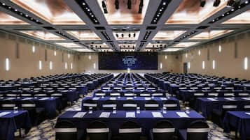 A large conference room with rows of tables and chairs, a stage with a screen displaying 'MAISON GLAD JEJO', and a modern ceiling design.