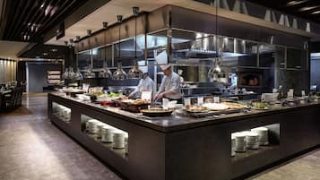 Una cocina moderna con chefs preparando alimentos, un bufé con diversos platos y un diseño limpio y contemporáneo.