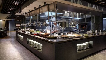 Una cocina moderna con chefs preparando alimentos, un bufé con diversos platos y un diseño limpio y contemporáneo.