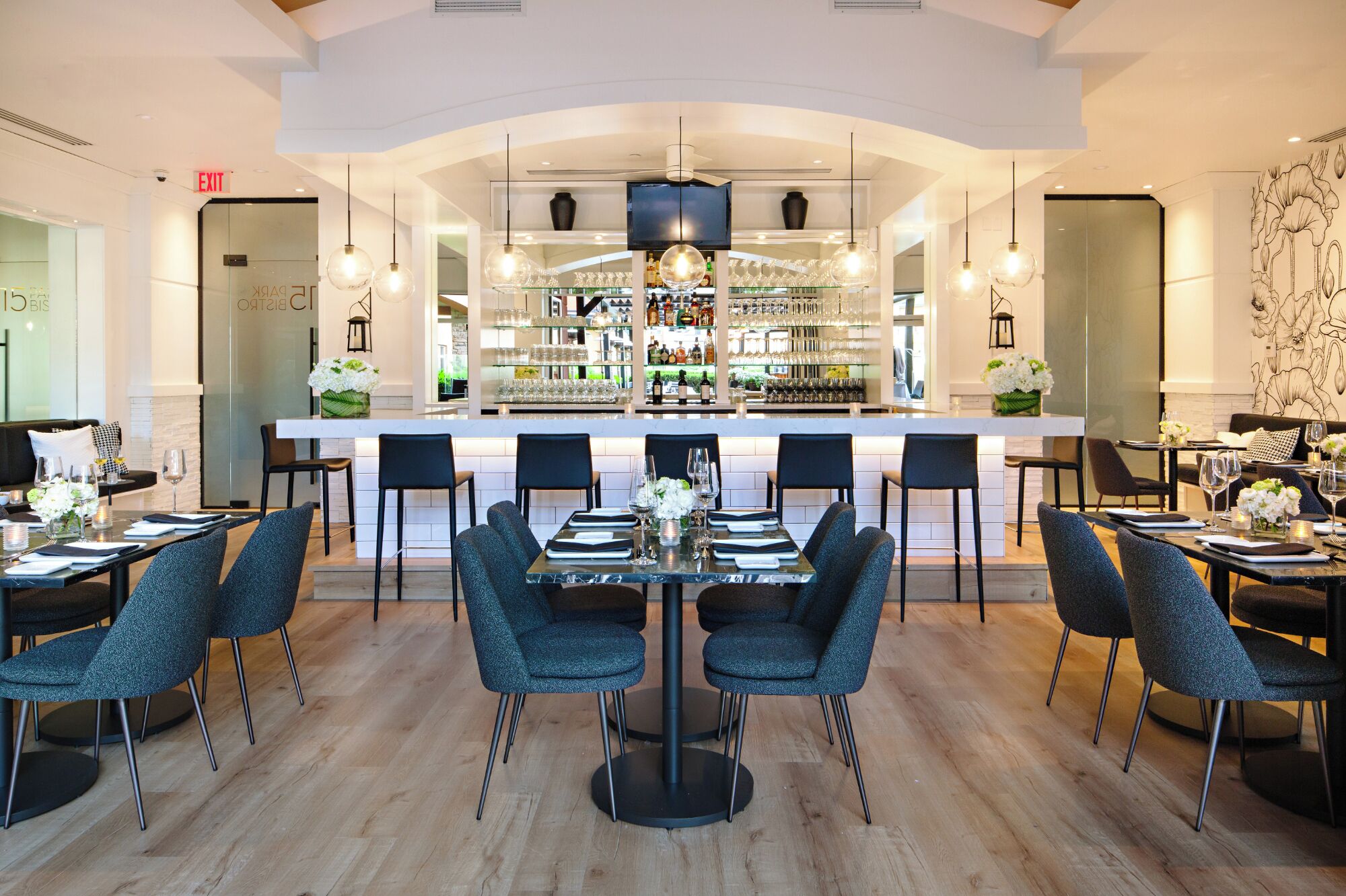 A modern restaurant with a central bar, black chairs, and a white table with a vase of flowers.