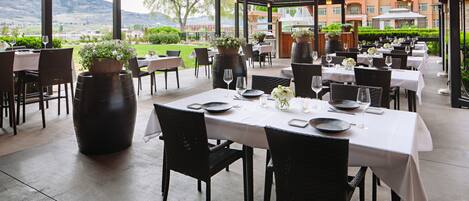 An outdoor dining area with tables set for two, potted plants, and a view of the landscape.
