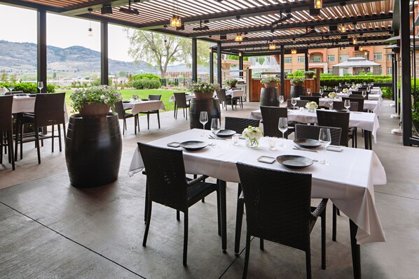An outdoor dining area with tables set for two, potted plants, and a view of the landscape.
