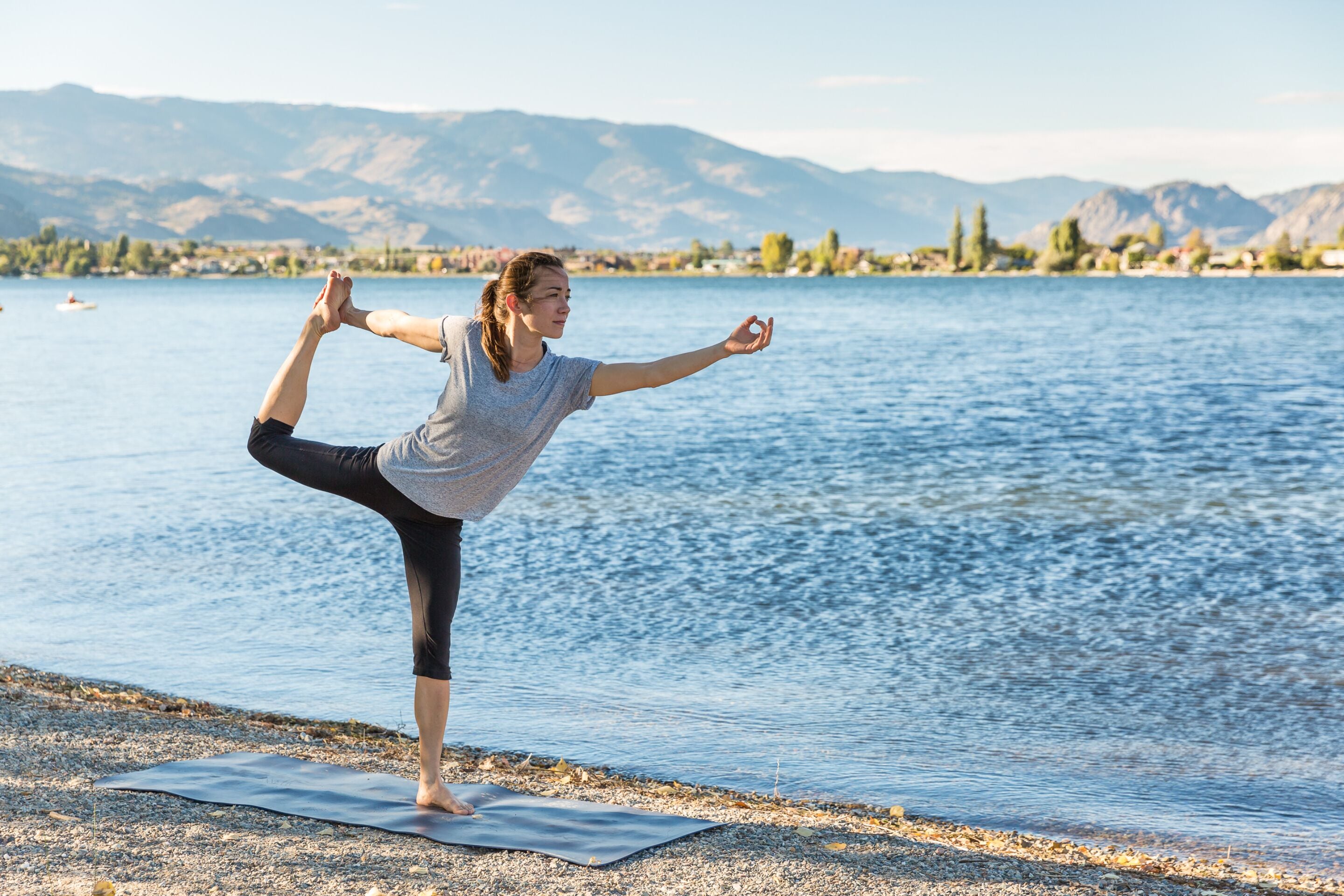 A person performing a yoga pose on a mat by a lake with mountains in the background.