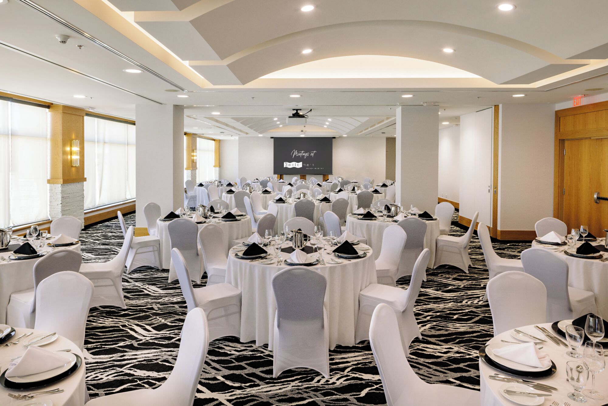 Een vergaderzaal met ronde tafels, witte stoelen en een projectiescherm.