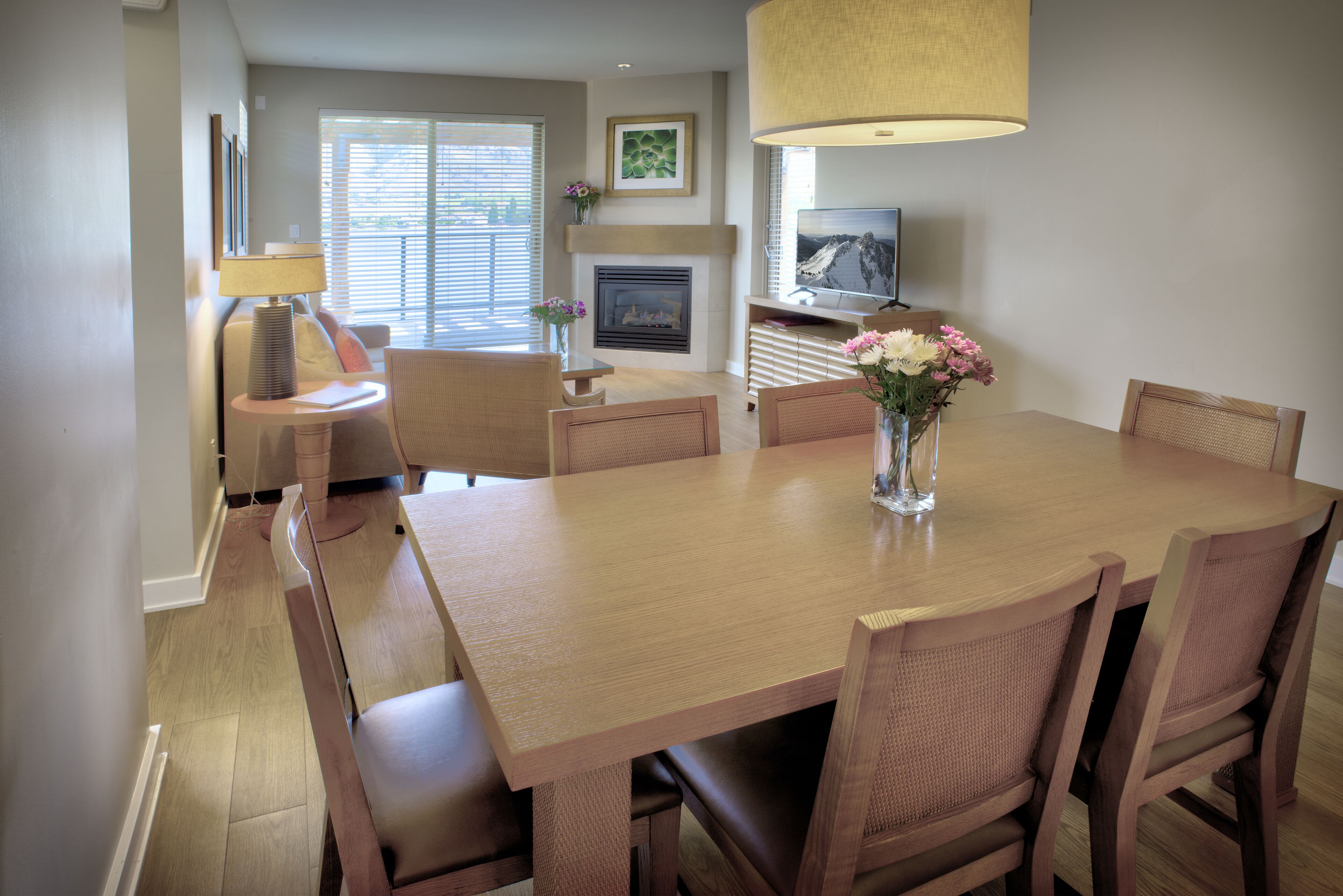 A dining area with a wooden table, wicker chairs, a vase with flowers, a fireplace, and a television on a stand.