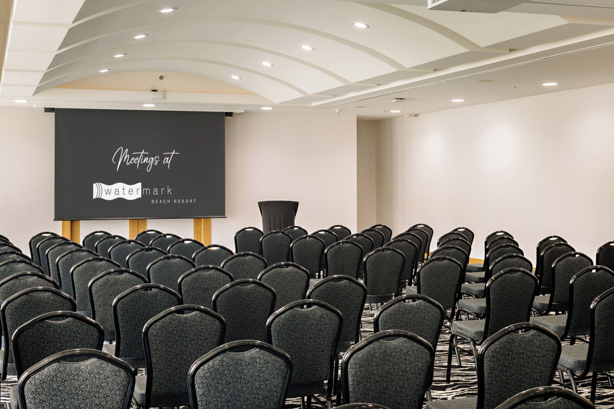 A conference room with rows of black chairs, a projector screen displaying "Meetings at Watermark Beach Resort", and a modern ceiling design.