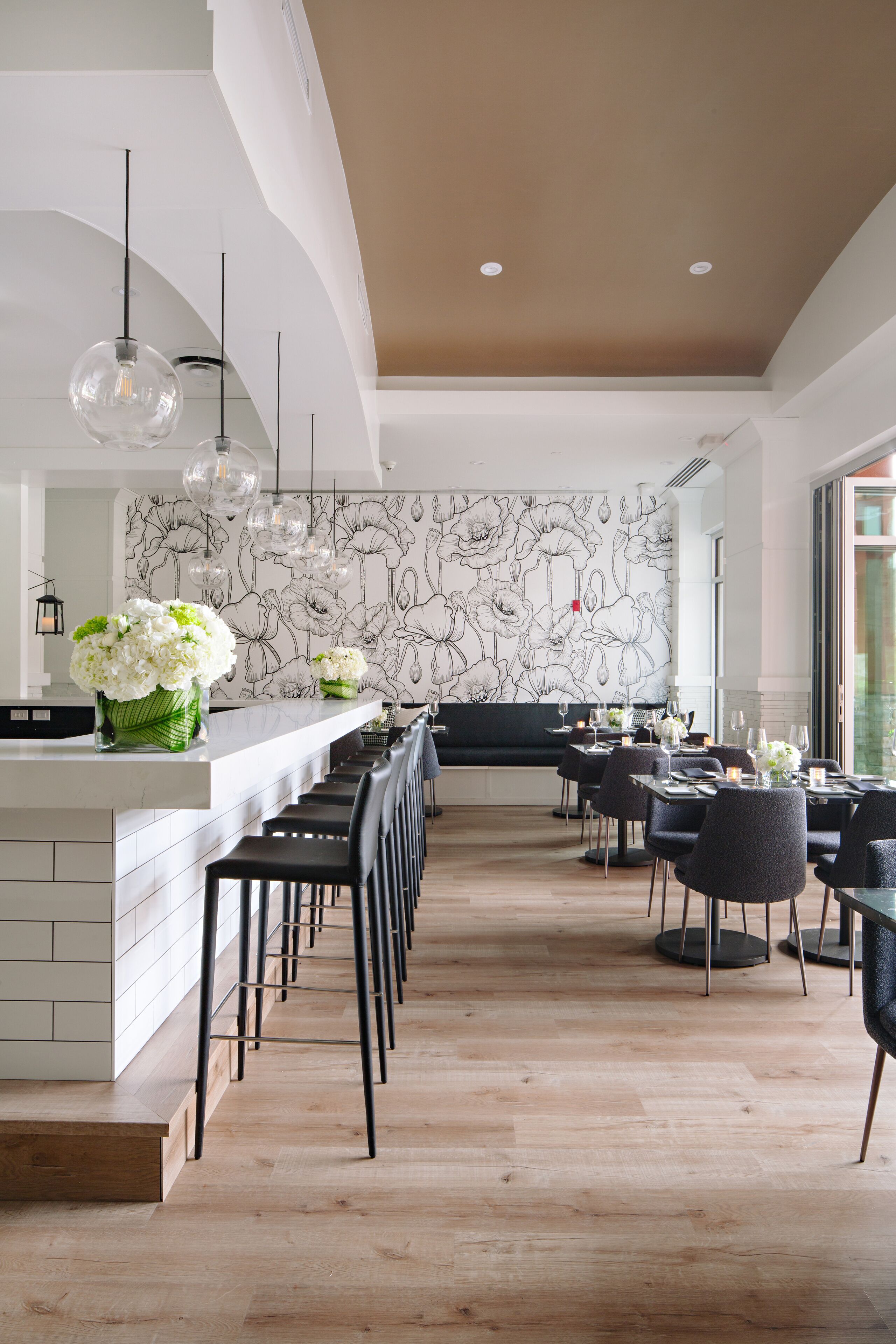 A modern restaurant with a long counter, black chairs, and a floral wall mural.
