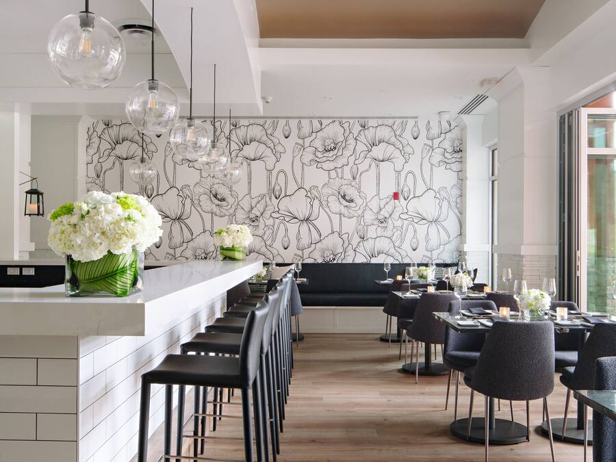 A modern restaurant with a long counter, black chairs, and a floral wall mural.