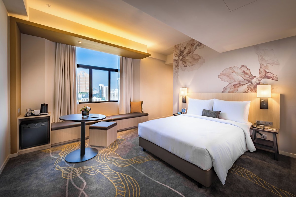 Ein Hotelzimmer mit Bett, Nachttischen, Schreibtisch, Stuhl, einem Fenster mit Vorhängen und Blick auf die Stadt.