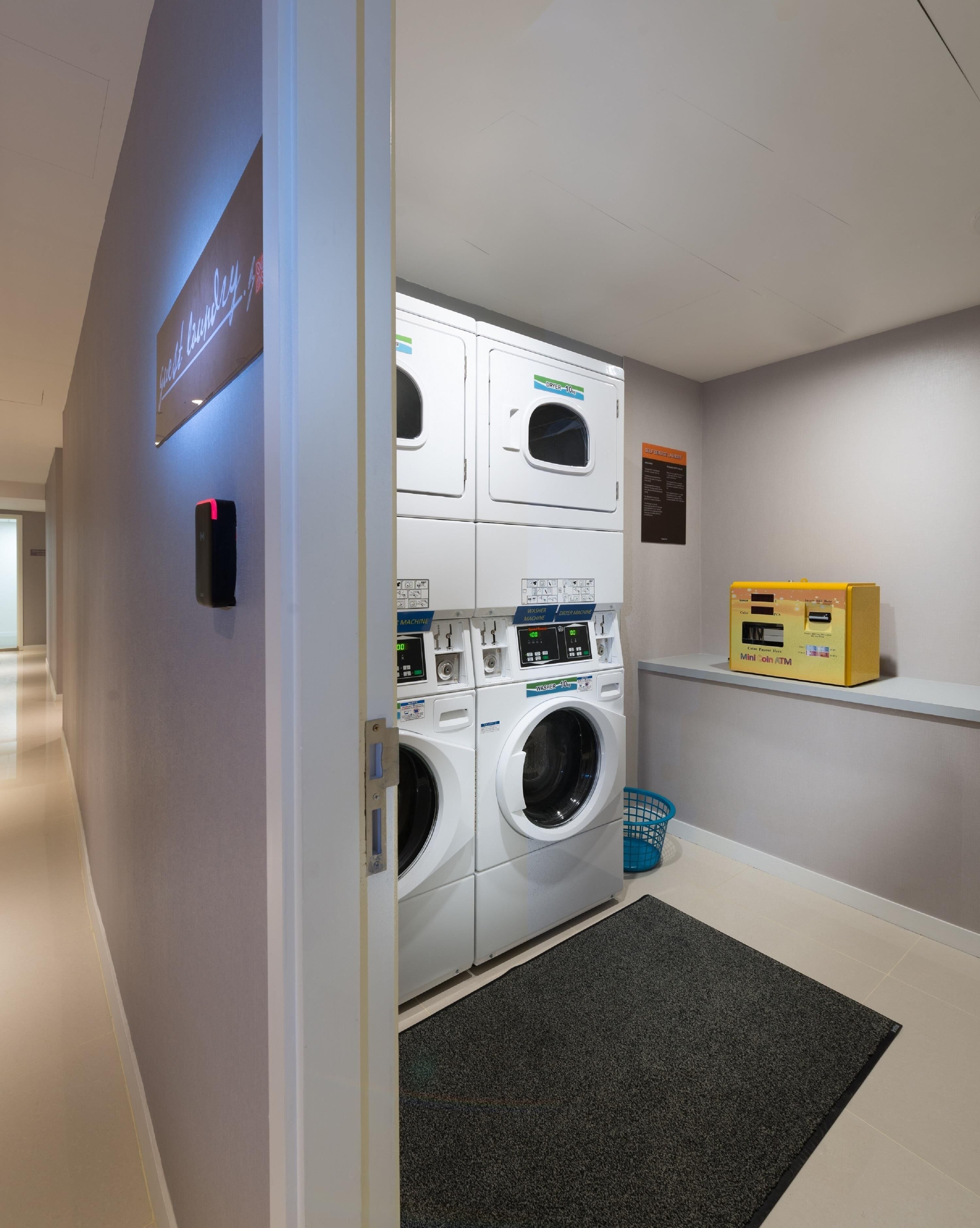 A laundry room with two stacked front-loading washers and dryers, a blue laundry basket, a mini coin ATM, and a sign indicating the room's name.