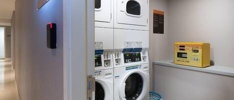 A laundry room with two stacked front-loading washers and dryers, a blue laundry basket, a mini coin ATM, and a sign indicating the room's name.