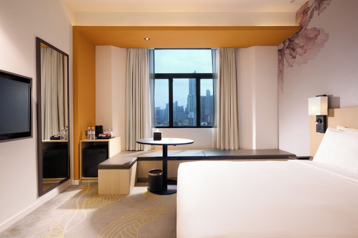 Ein Hotelzimmer mit Bett, Schreibtisch, Stuhl, Fernseher, einem Fenster mit Vorhängen und Blick auf die Stadt.