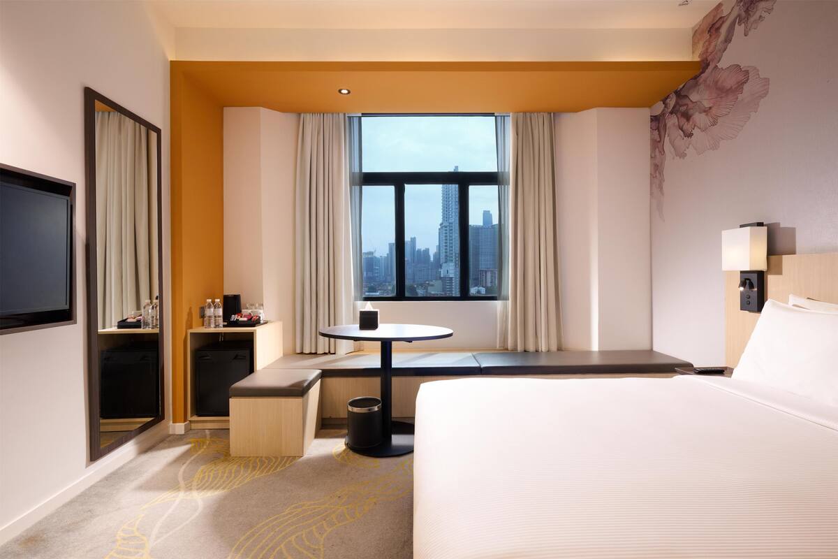 A hotel room with a bed, a desk, a chair, a TV, a window with a city view, and a trash can.