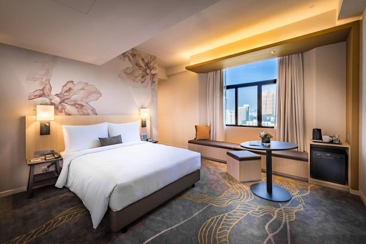 A hotel room with a large bed, bedside tables, a desk, a chair, a round table, a window with curtains, and a city view.