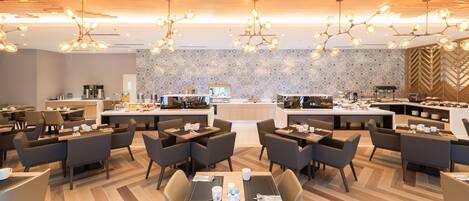 Una sala da pranzo moderna in un hotel, con diversi tavoli, sedie e una zona cucina centrale.