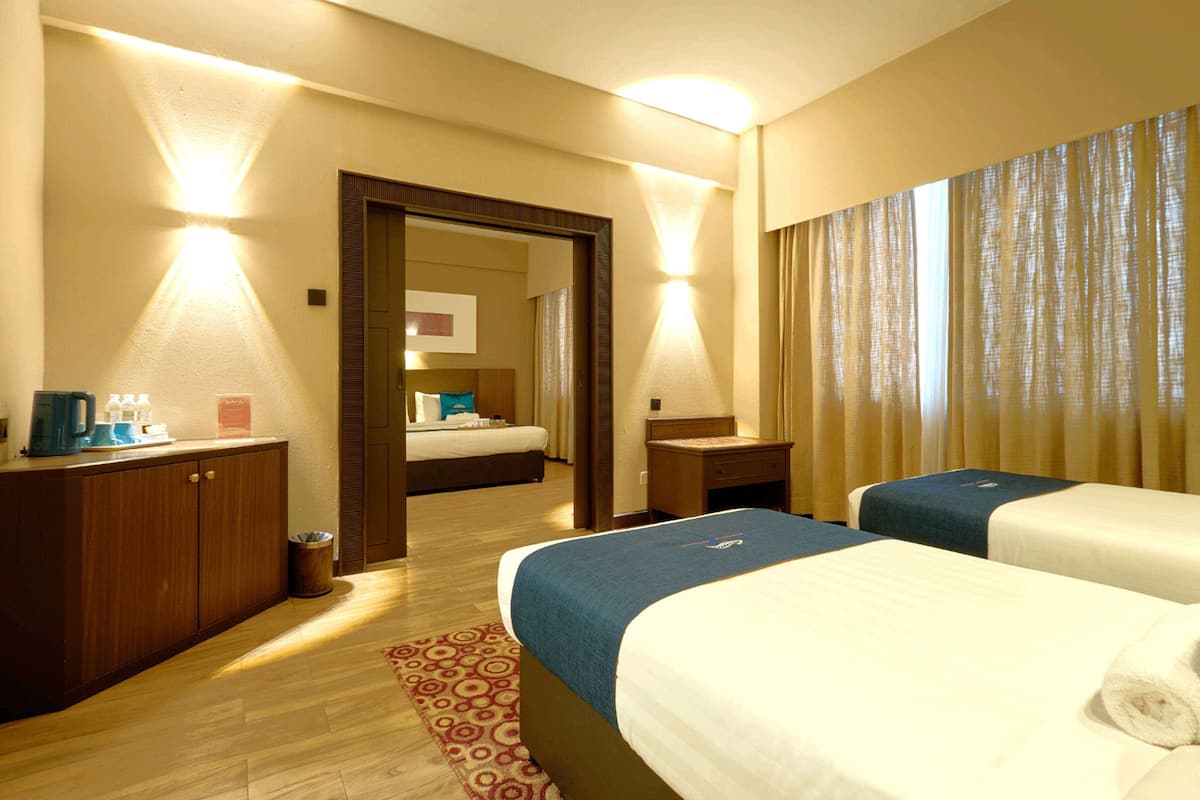 A hotel room with two beds, a wooden wardrobe, a bedside table, a lamp, and a mirror.
