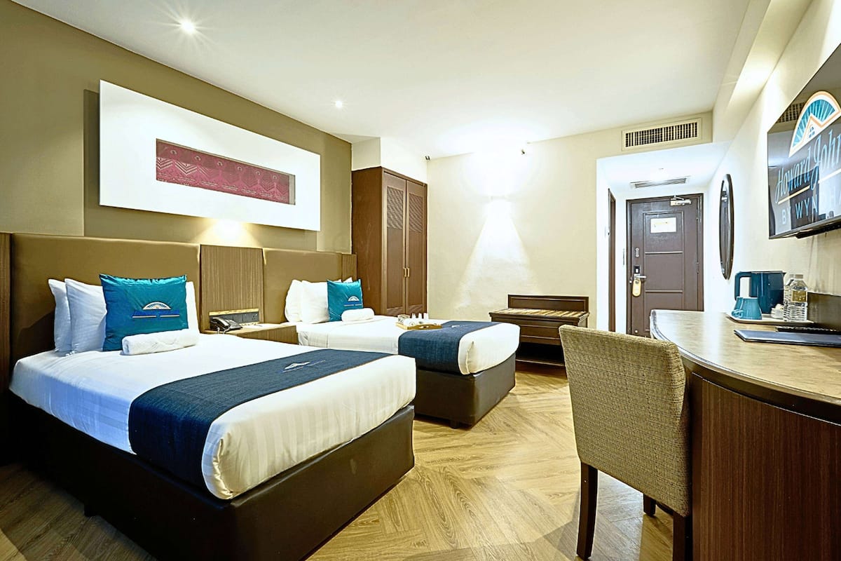 A hotel room with two beds, a desk, and a chair. There is a large window, a door, and a framed picture on the wall.