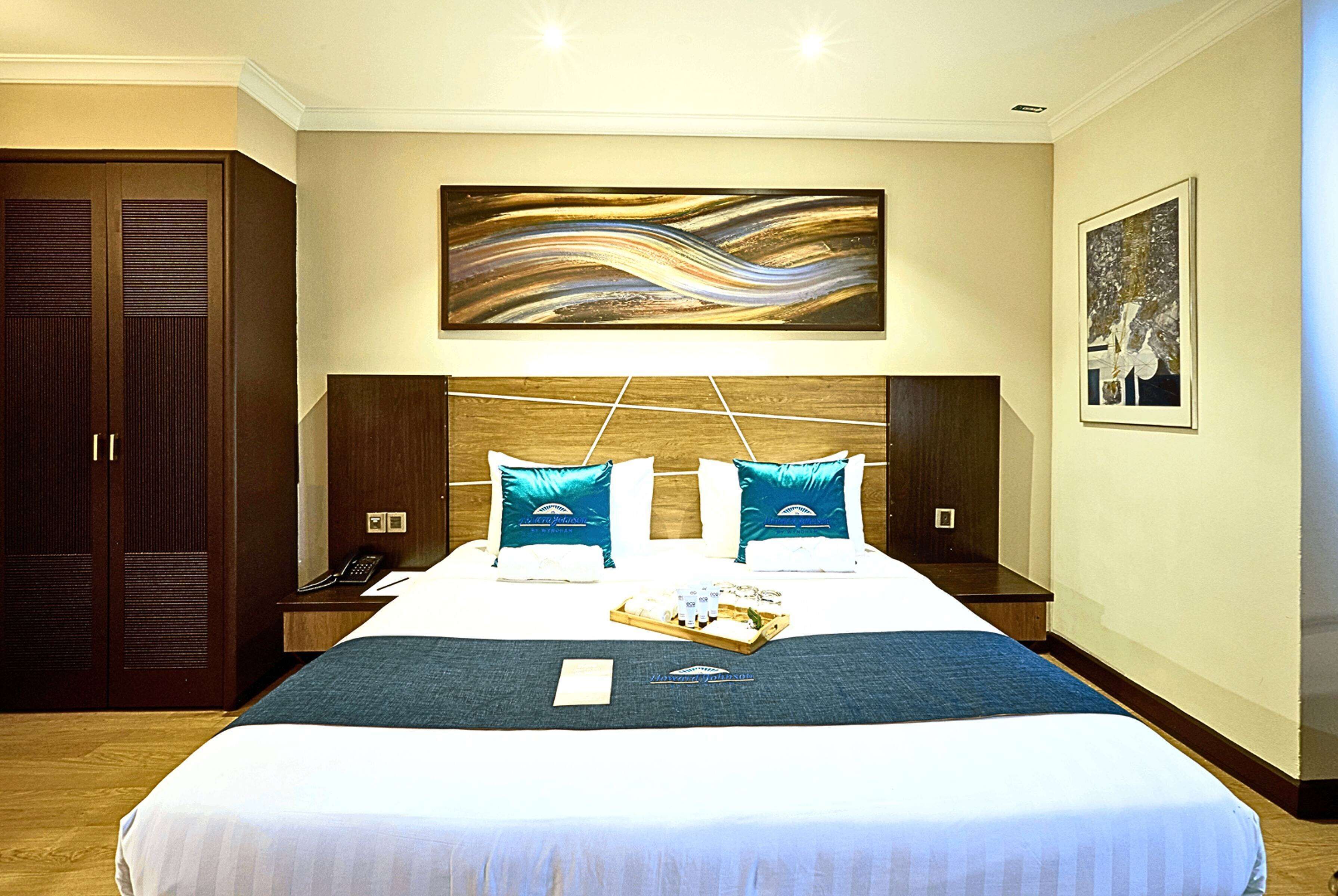 Une chambre d’hôtel avec un grand lit, deux tables de chevet, une tête de lit en bois, un tableau au-dessus du lit et une armoire.