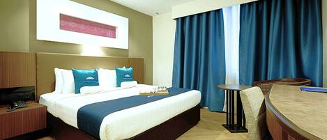 A hotel room with a large bed, a desk with a chair, a television, and a window with curtains.