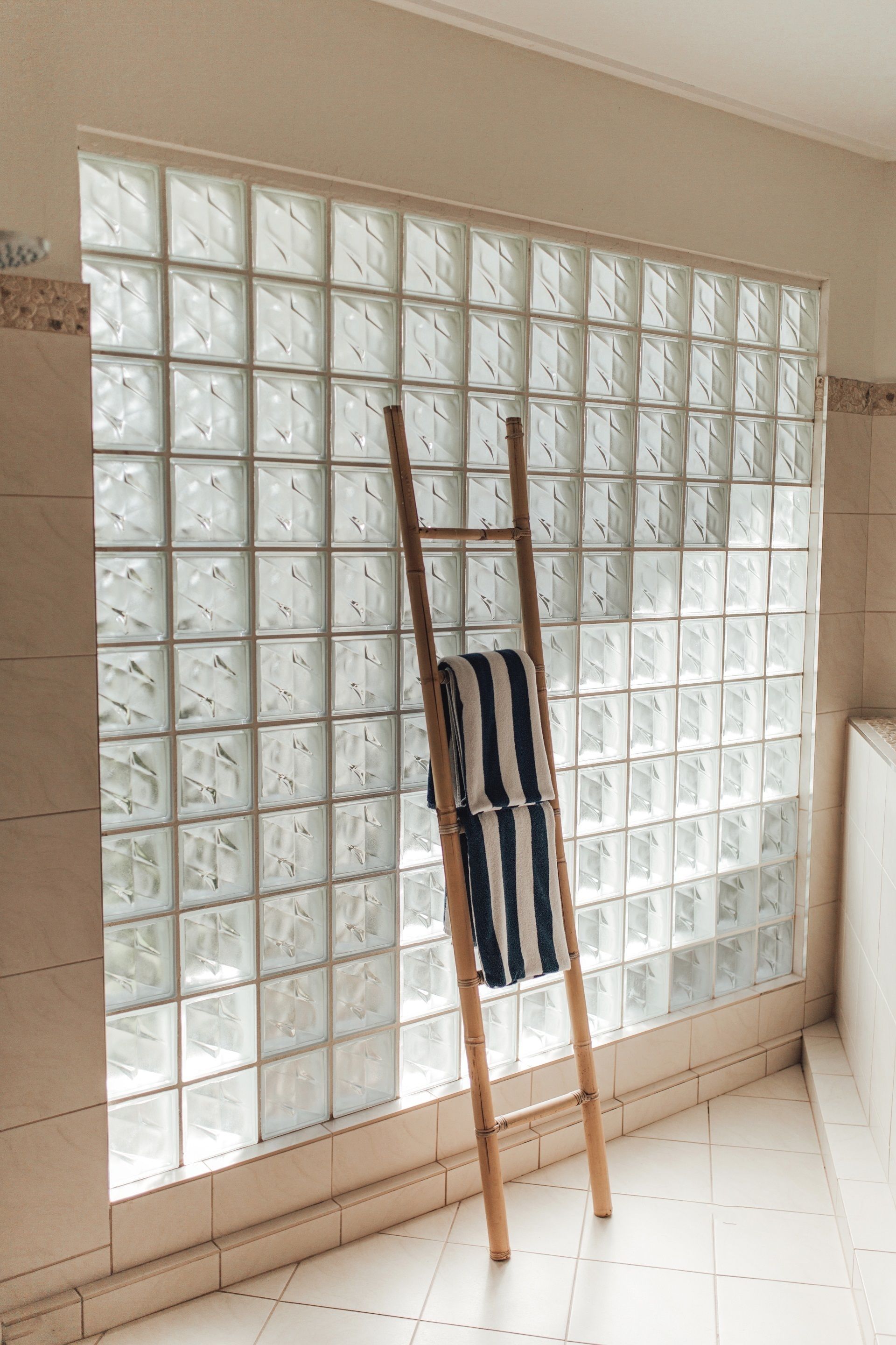 Una habitación con un ventanal grande cubierto por paneles de vidrio esmerilado, una escalera de madera apoyada en la pared y una toalla a rayas colgada de la escalera.