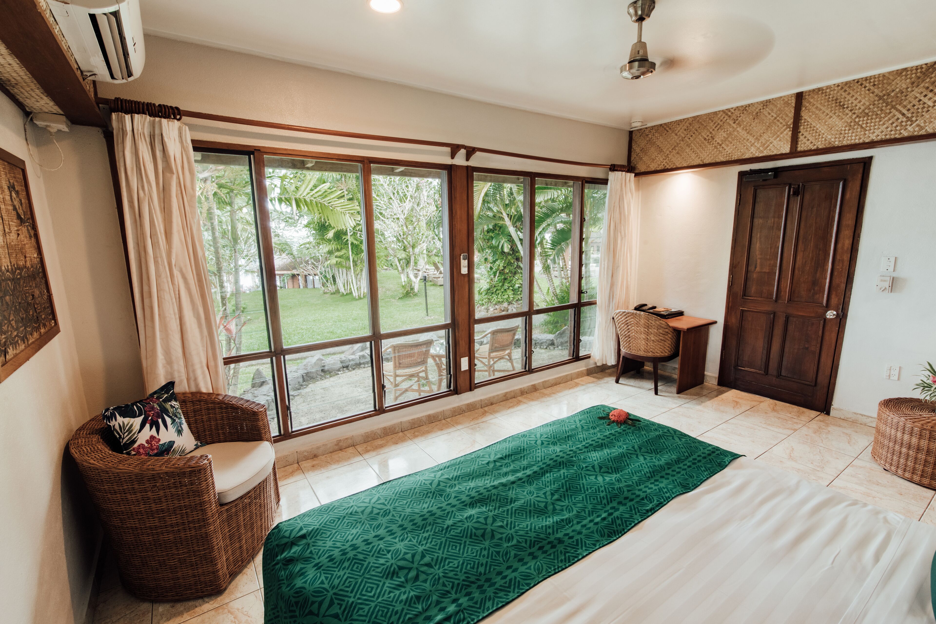 Una habitación con un gran ventanal corredero, una silla de mimbre, una cama con edredón estampado en verde y una puerta de madera.
