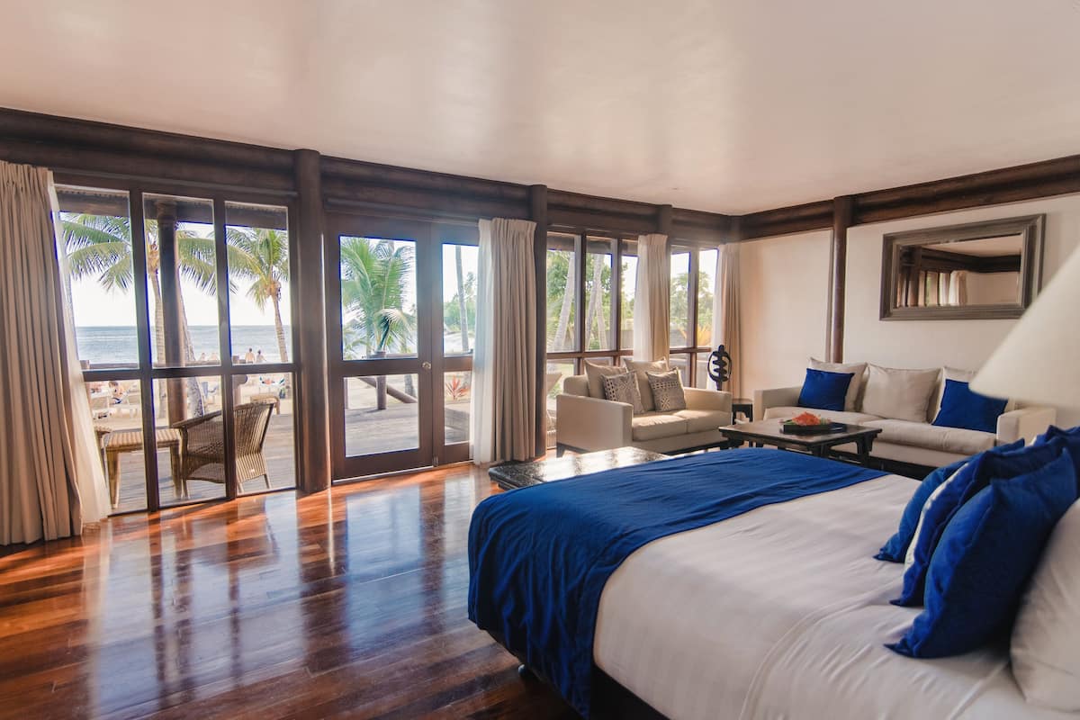 Un dormitorio amplio con una cama grande, un sofá y una mesa de comedor. Hay una puerta corrediza de vidrio que da a un balcón con vista a palmeras y al océano.