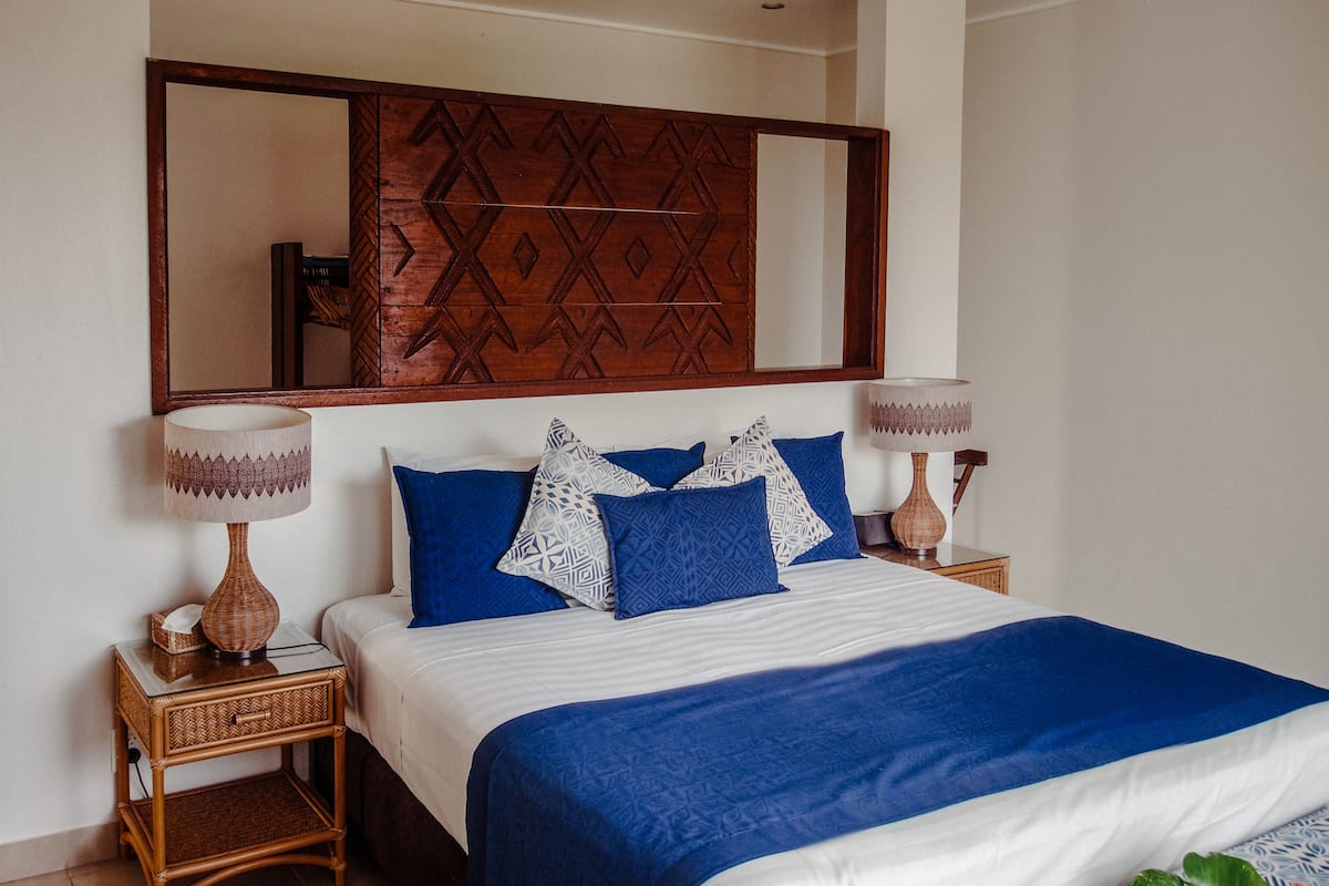 Una cama bien hecha con ropa de cama azul y blanca, dos mesitas de noche con lámparas y un espejo grande sobre la cama.