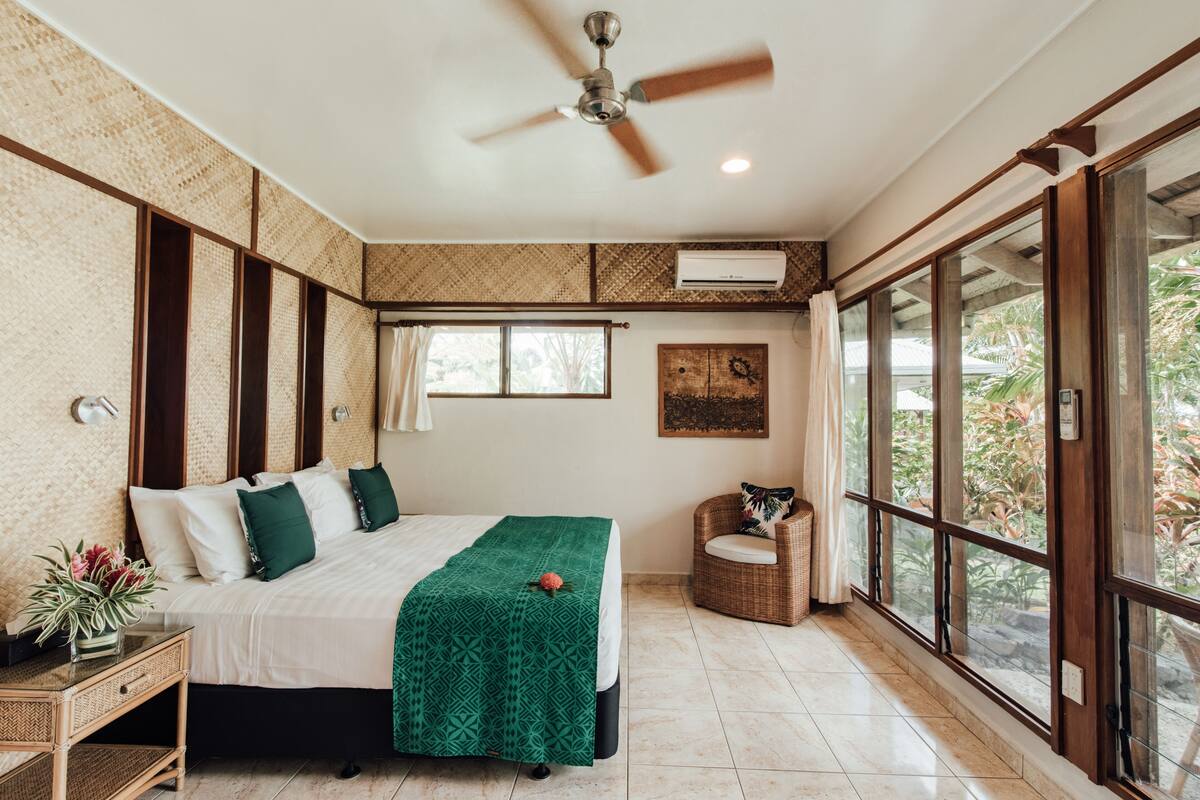 Un dormitorio con cama, ventilador de techo, ventana y vistas a la vegetación exterior.