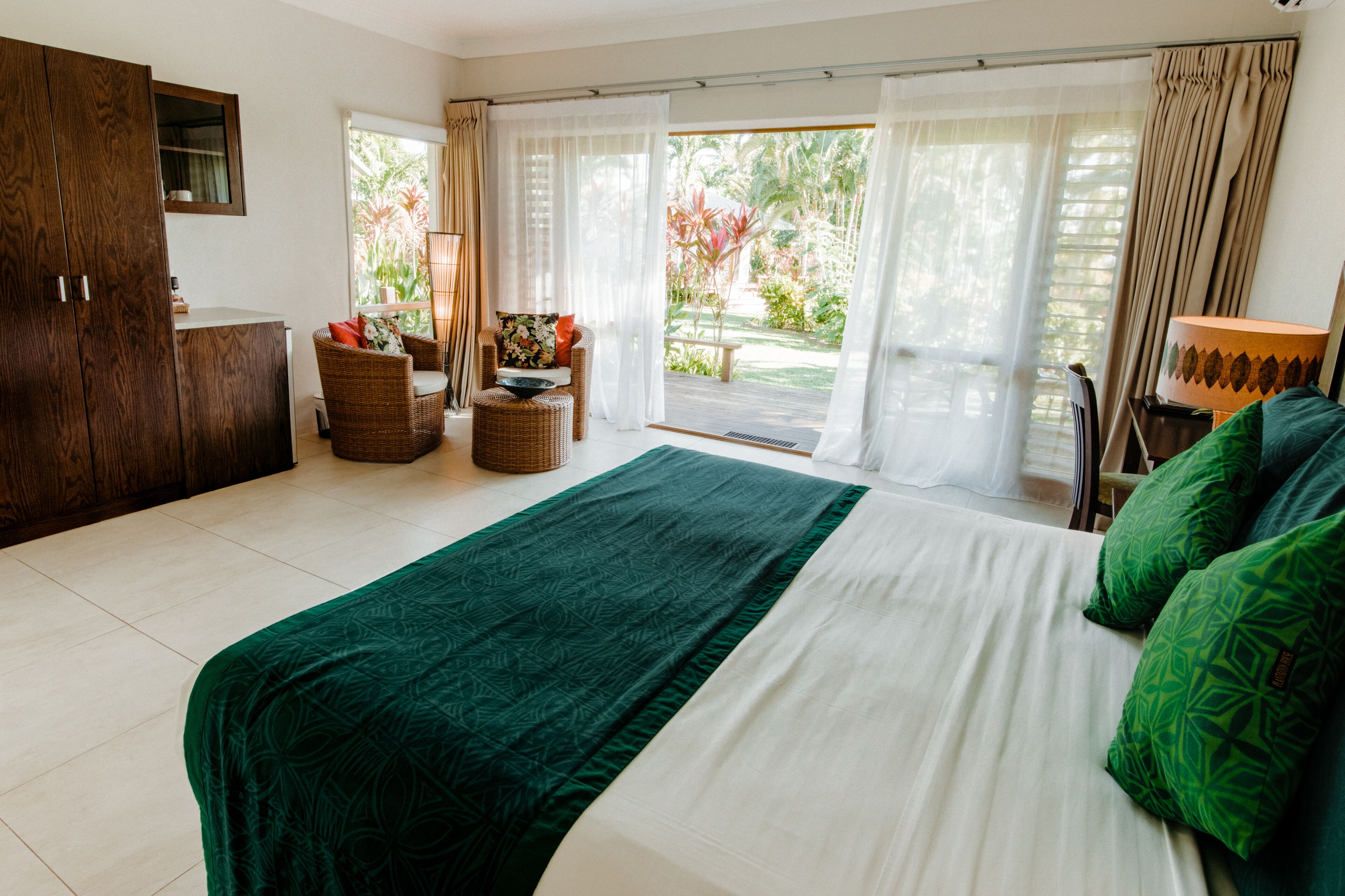 A bedroom with a large bed, a wooden wardrobe, a wicker chair, a small table with a bowl, and a view of the outdoors through large windows.