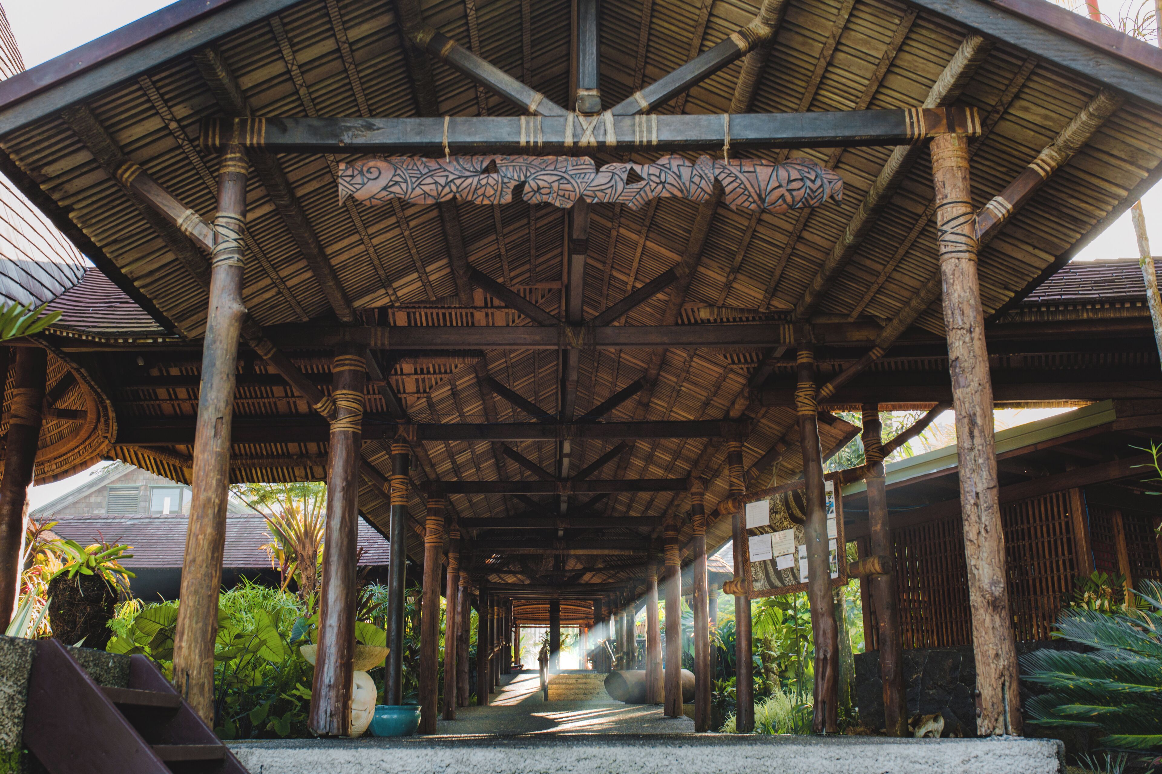 A covered walkway with wooden pillars and a patterned ceiling, surrounded by greenery.