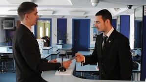 Two men in suits at a reception desk in a modern hotel lobby with blue accents and a ceiling-mounted security camera.