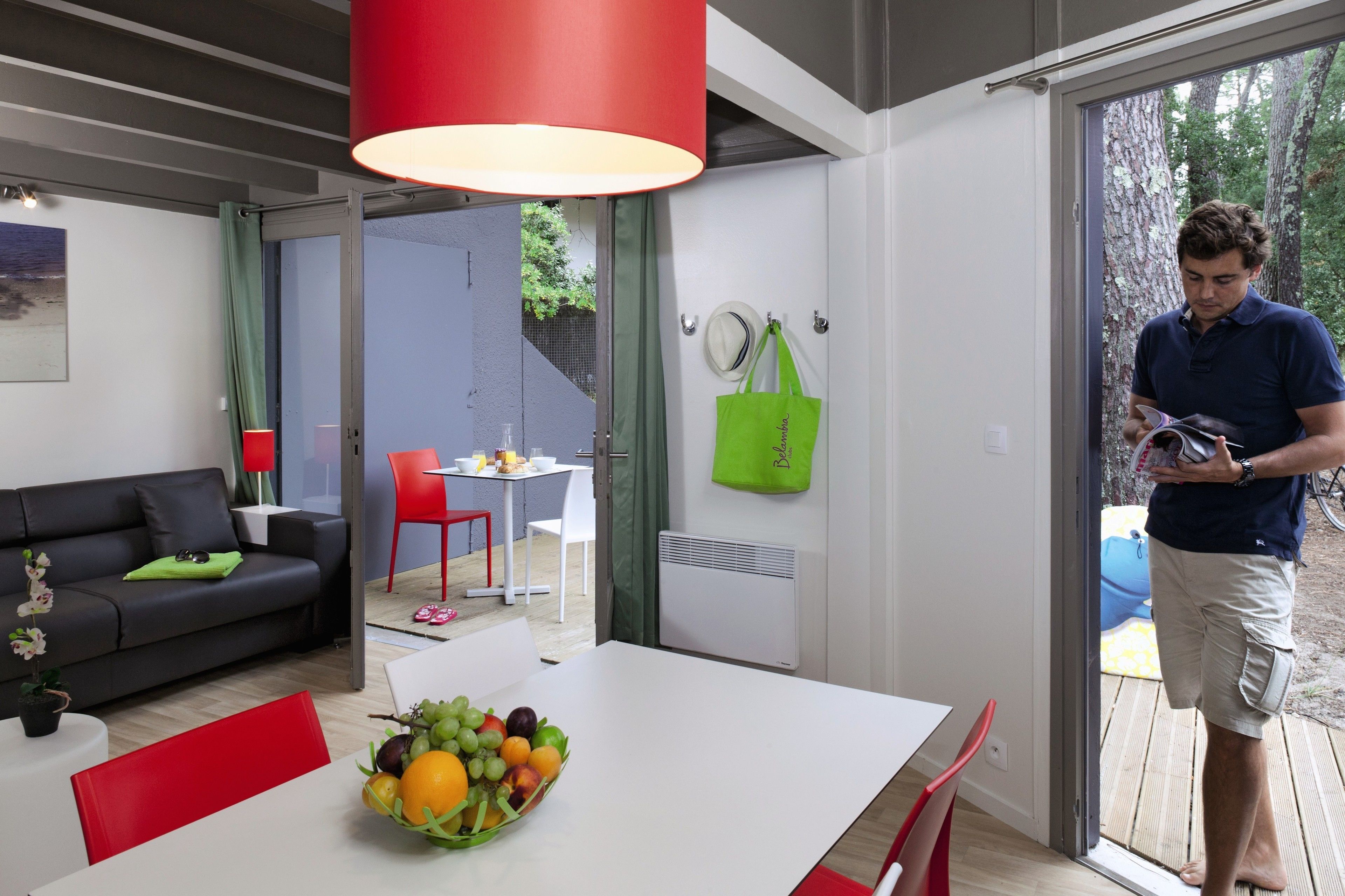 A modern living room with a white dining table, red chairs, a black sofa, and a fruit bowl.