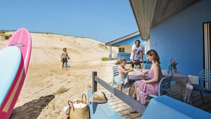 Un patio frente a la playa con terraza de madera, una mesa y sillas. Se ven sombrillas y una tabla de surf.