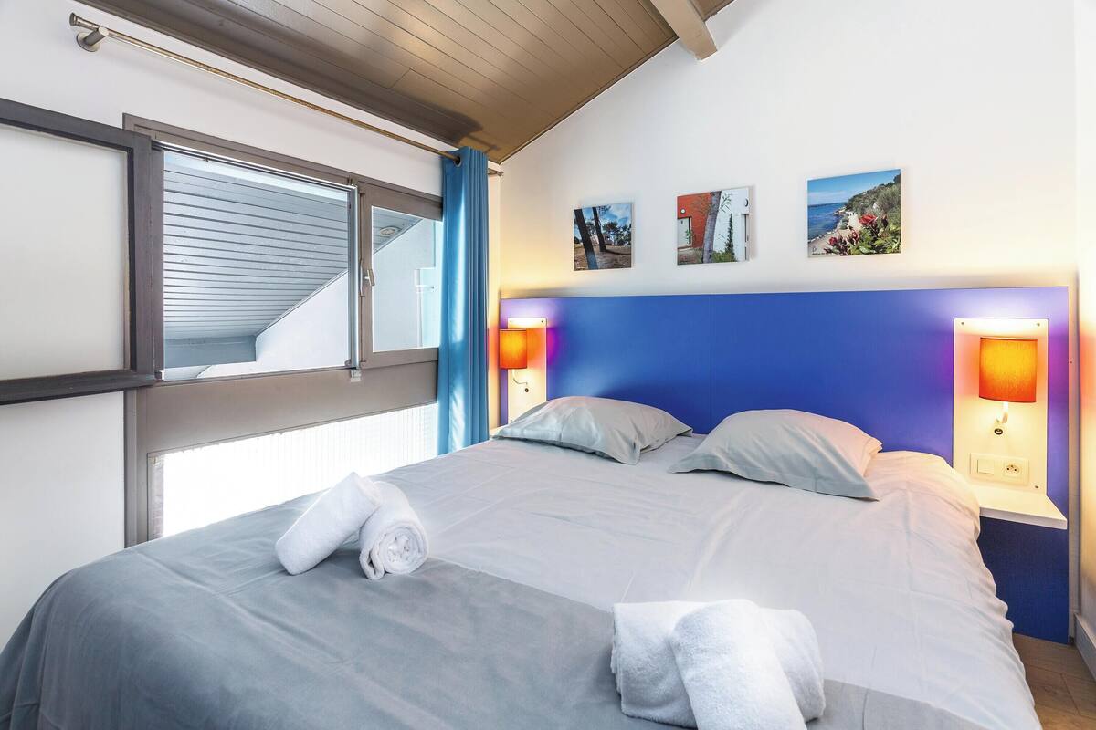 Una habitación de hotel con una cama, dos lámparas de noche, un ventanal con persianas y tres cuadros en la pared.