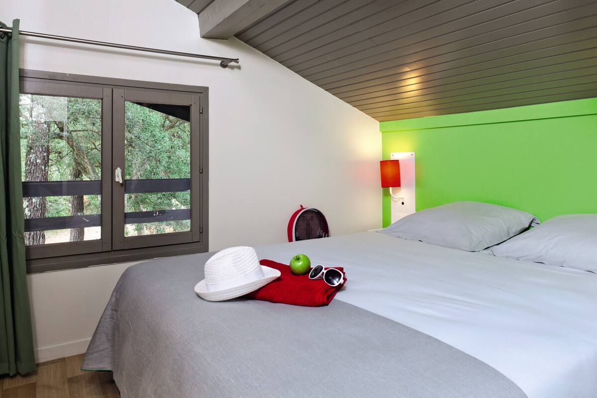 Un dormitorio con cama, ventana con vista a árboles, pared verde y toalla roja con sombrero y gafas de sol.