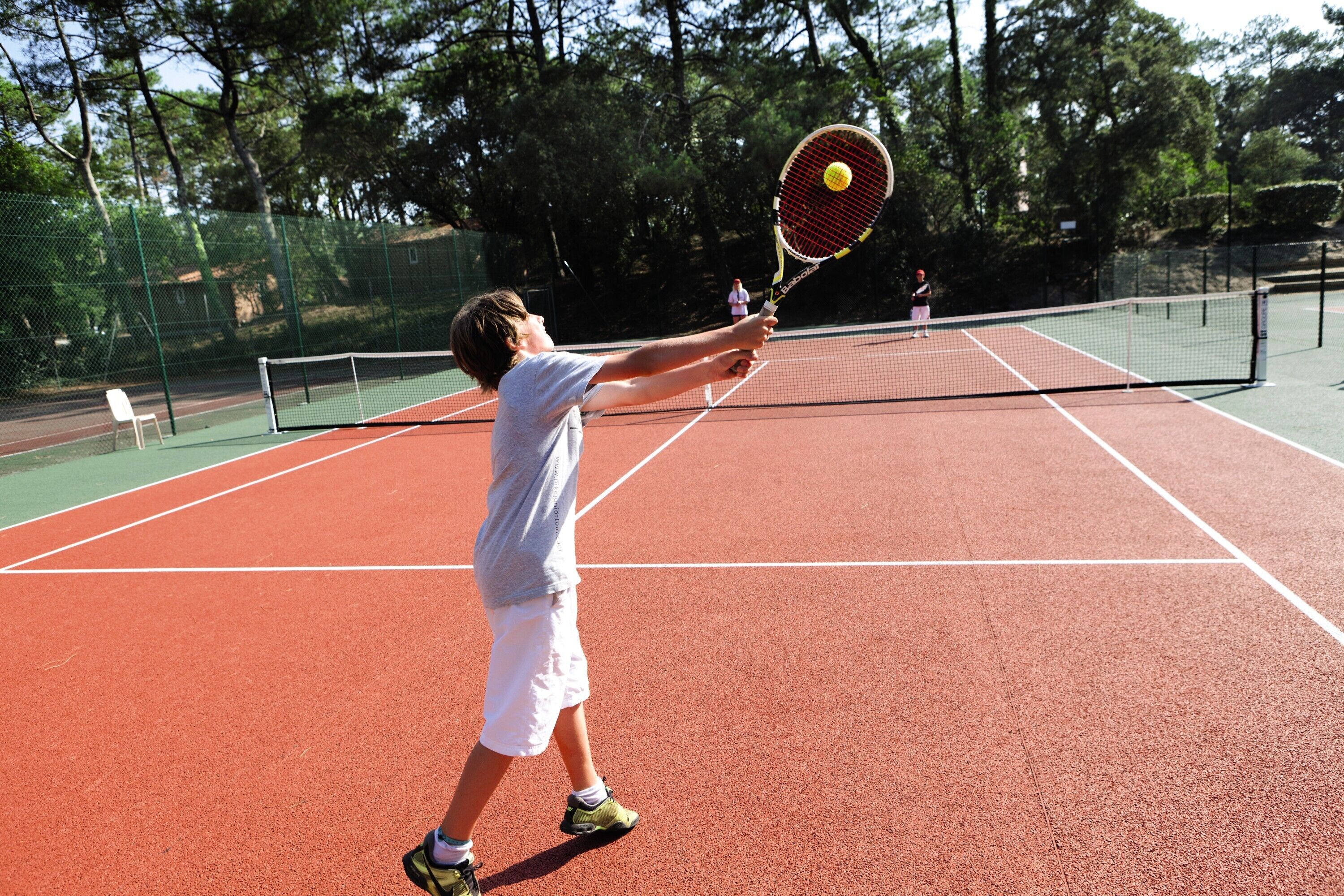 A tennis court with a net, a player in a white shirt and shorts, and a tennis ball in motion.