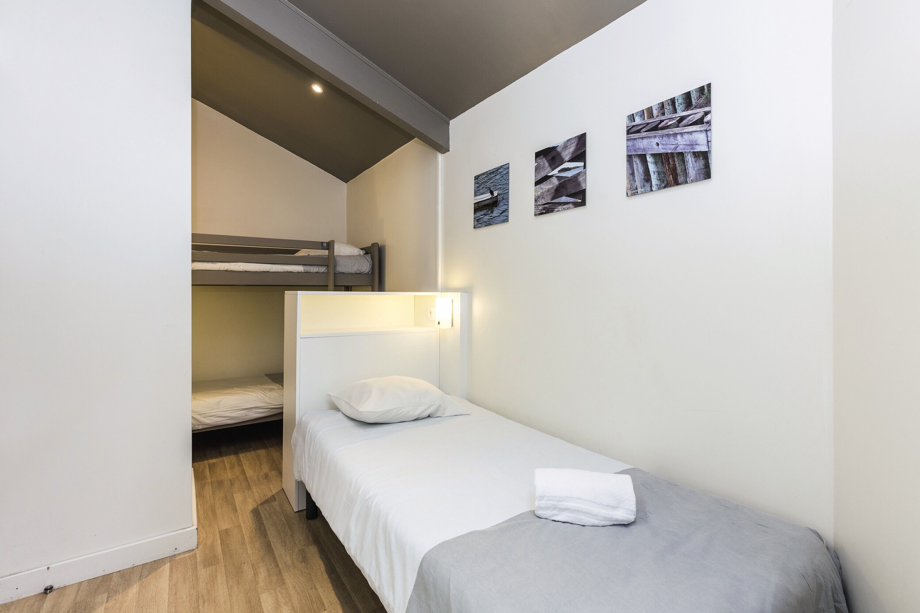 Una habitación pequeña con una cama individual y una litera al fondo, una mesita de noche y tres cuadros colgados en la pared.