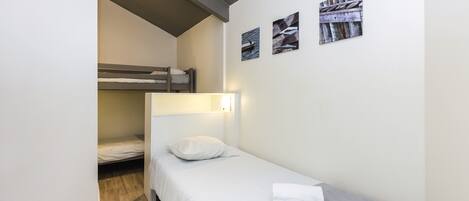 Una habitación pequeña con una cama individual y una litera al fondo, una mesita de noche y tres cuadros colgados en la pared.