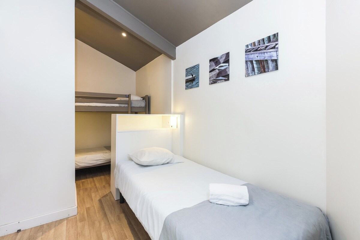 Una habitación pequeña con una cama individual y una litera al fondo, una mesita de noche y obras de arte colgadas en la pared.