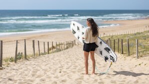 Una persona sosteniendo una tabla de surf en una playa arenosa con una cerca y el océano al fondo.