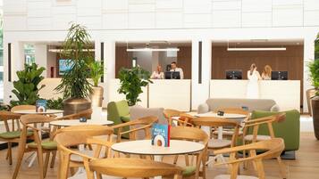 Een hotelhal met ronde tafels en stoelen, receptiebalies en potplanten.