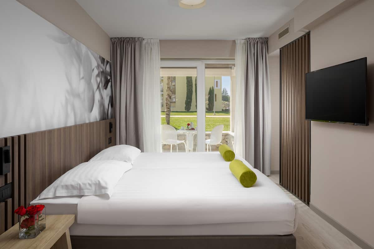 Ein Hotelzimmer mit Bett, Fernseher, Fenster mit Vorhängen, Blick ins Freie und einem Tisch mit Blumenvase.