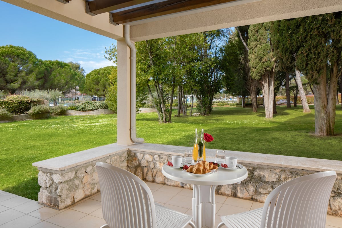 Eine Terrasse mit einer Steinsitzbank und zwei Stühlen, einem Tisch mit Frühstücksangeboten und Blick auf eine gepflegte Rasenfläche und Bäume.